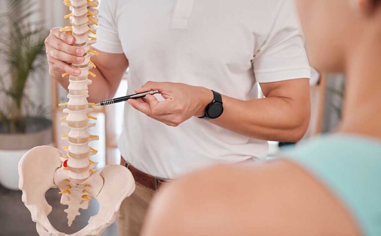 A chiropractor explains spinal anatomy to an adult patient using a model of the human spine in a clinical setting.